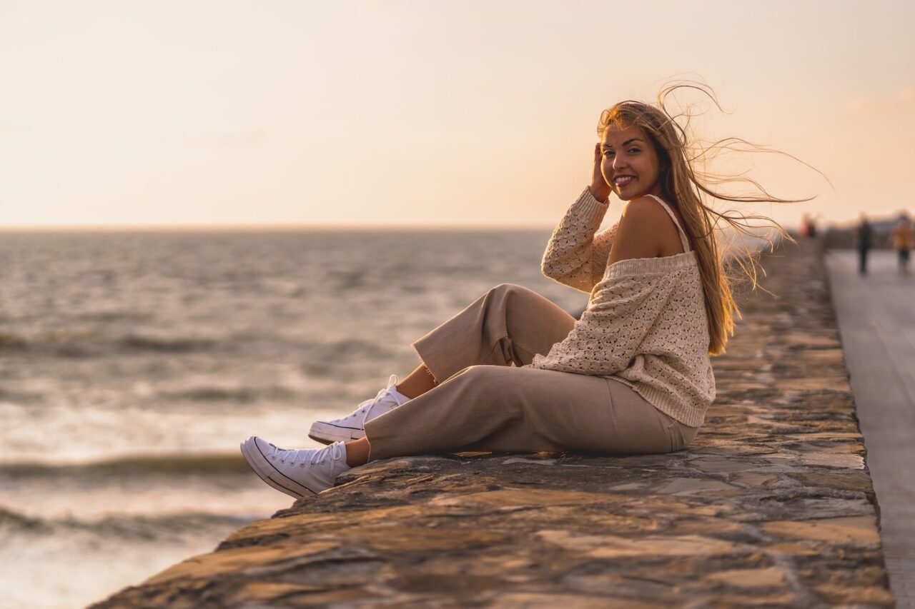 Portrait of a young blonde Caucasian woman sitting by the sea in a white short shirt